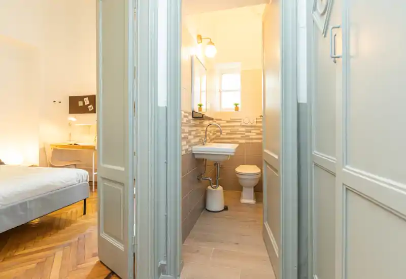Camera da letto luminosa e moderna con letto matrimoniale visibile, pavimento in parquet e una scrivania; il bagno en-suite è visibile attraverso le porte aperte.