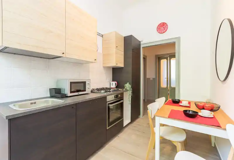 Cucina moderna compatta con basi scure e pensili in legno chiaro, forno incassato e piano cottura a gas, più un piccolo tavolo da pranzo apparecchiato per due — luminosa, funzionale e ben curata.
