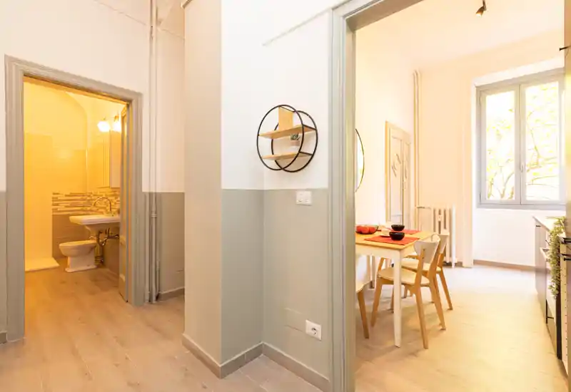Piccolo angolo pranzo luminoso visto attraverso una porta aperta, con tavolo e sedie in legno semplici, luce naturale da una grande finestra e arredamento minimal e caldo.