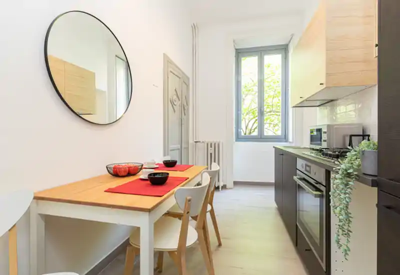 Cucina moderna compatta con forno integrato e piano a gas, piccolo tavolo in legno apparecchiato con tovagliette rosse e finestra luminosa che dona luce naturale.
