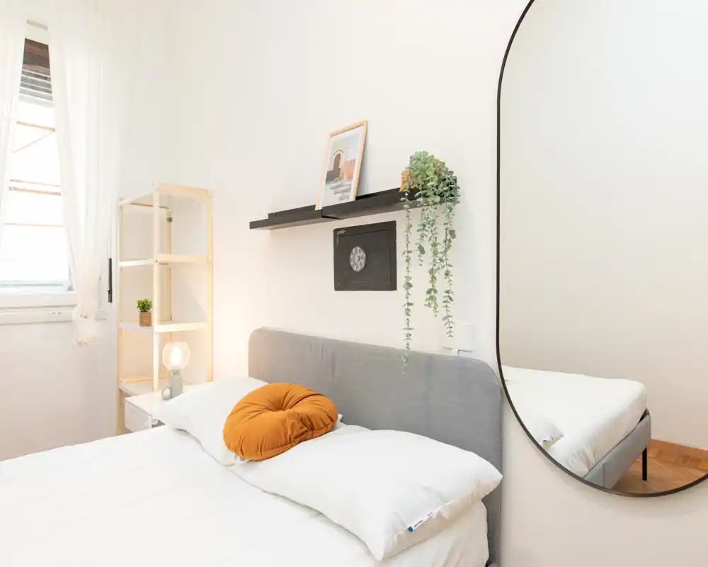 Camera da letto luminosa e minimale con testiera imbottita grigia, biancheria bianca e un cuscino rotondo ocra; luce naturale e mensole curate creano un ambiente tranquillo e accogliente.