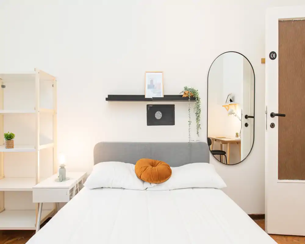 Camera da letto luminosa e minimal con letto matrimoniale al centro, biancheria bianca e un cuscino accentato caldo. Mensole pulite, lampada da comodino e uno specchio ovale a parete aggiungono stile funzionale adatto a un annuncio.