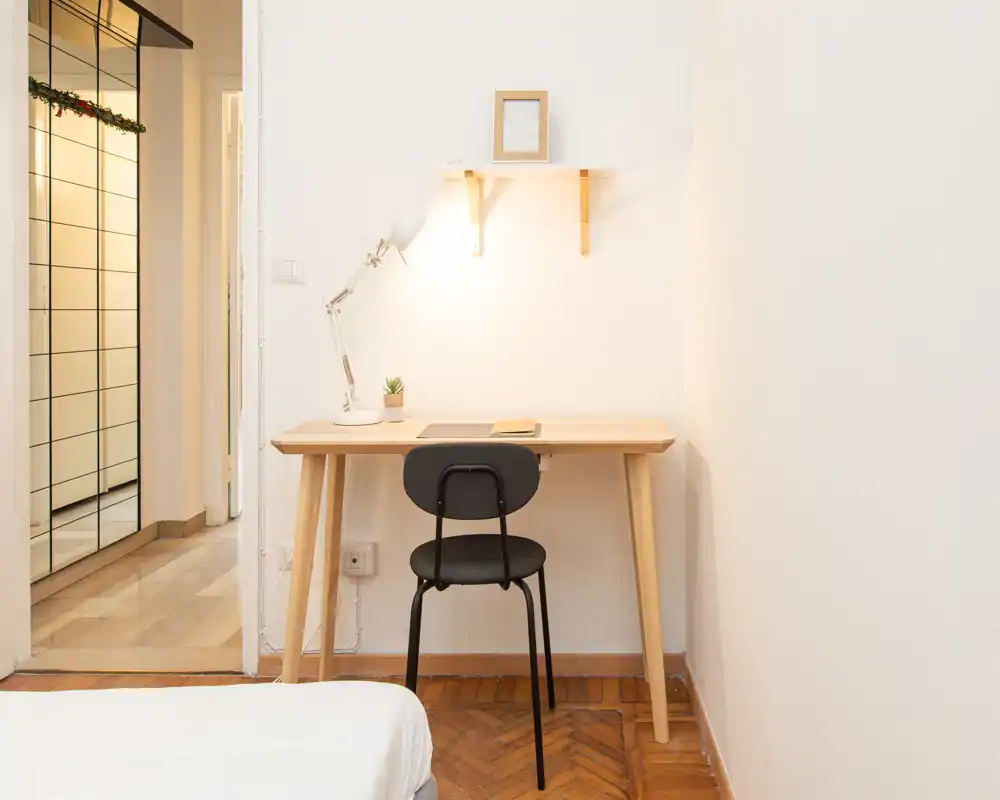 Angolo camera luminoso e minimale con una piccola scrivania in legno, lampada da tavolo e sedia accanto al letto. Stile pulito di ispirazione nordica e illuminazione calda creano un ambiente tranquillo per lavorare.