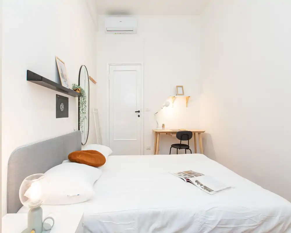 Camera da letto luminosa e minimal con letto matrimoniale ben rifatto, lampada da comodino e un piccolo angolo studio: ambiente curato e accogliente, ideale per un annuncio.