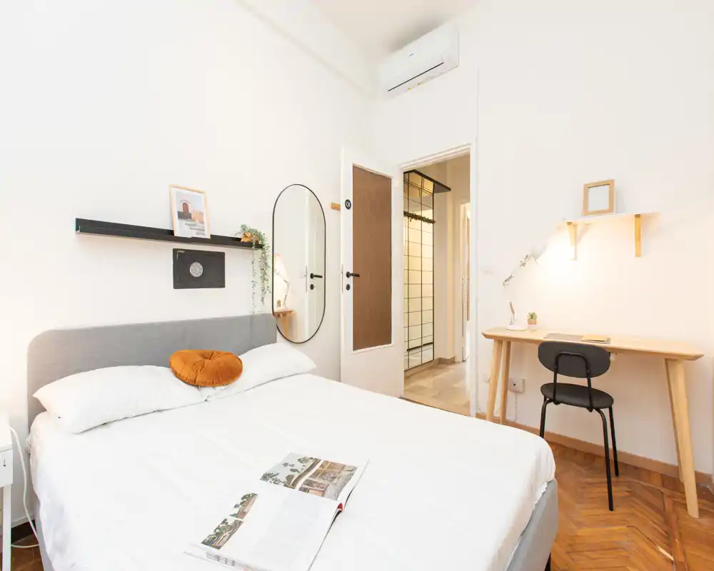 Camera da letto luminosa e moderna con letto matrimoniale ben rifatto, scrivania minimal e specchio a parete — perfetta per un soggiorno confortevole.