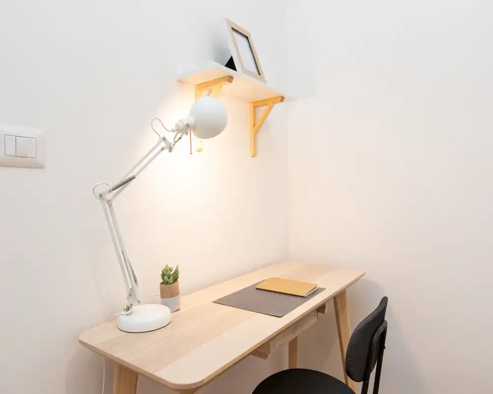 Angolo studio minimale e ben illuminato con scrivania in legno chiaro, lampada da tavolo regolabile, una piantina e sedia semplice — un angolo compatto e ordinato per lavorare.