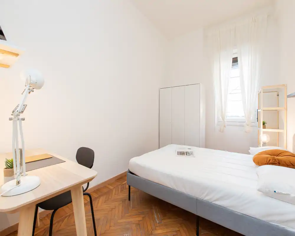 Camera da letto luminosa e minimale con letto matrimoniale, scaffalatura a fianco del letto, armadio e una scrivania vicino alla finestra. Il parquet in legno e la luce naturale donano un’atmosfera calda e accogliente, perfetta per affitti brevi.