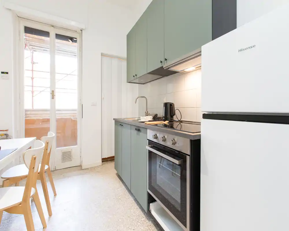 Angolo cottura moderno e luminoso con pensili verdi, forno integrato, piano cottura e frigorifero bianco. Un piccolo tavolo e la porta verso il balcone assicurano luce naturale e atmosfera accogliente.