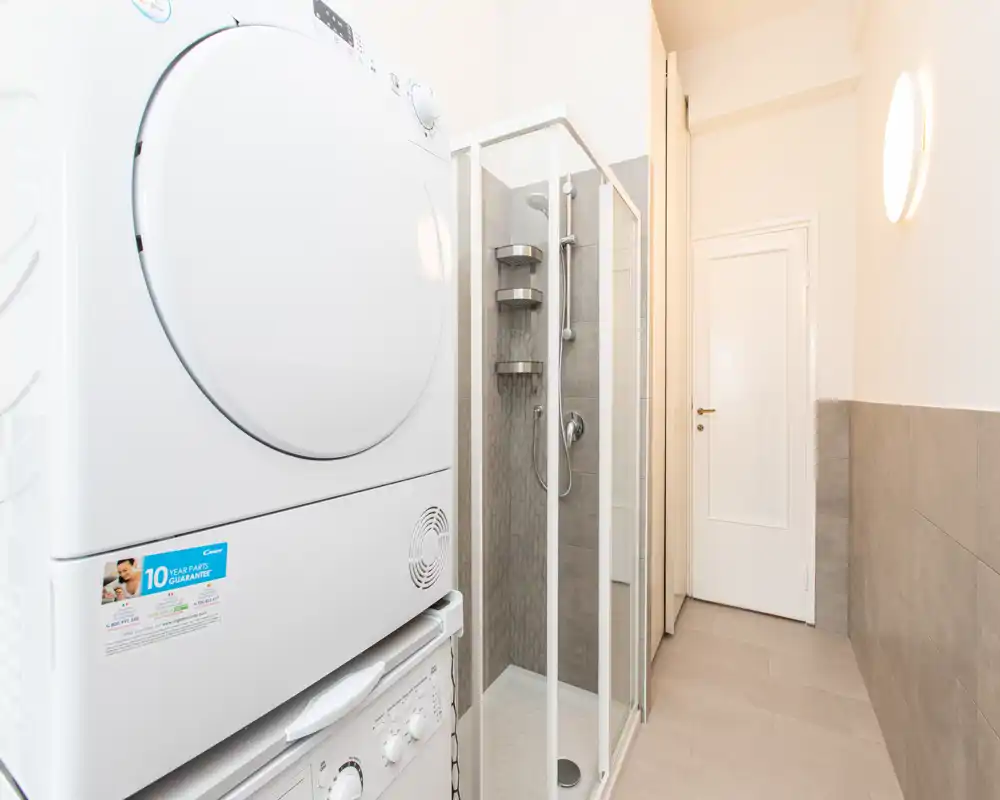 Bagno moderno e compatto con cabina doccia in vetro e colonna lavatrice/asciugatrice; piastrelle neutre e illuminazione luminosa creano uno spazio funzionale e ben tenuto.