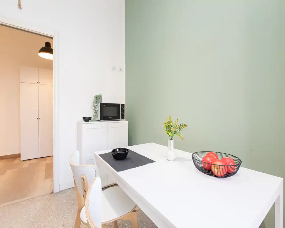Zona pranzo luminosa e minimal con tavolo bianco, due sedie e una ciotola di frutta decorativa su una parete verde tenue — perfetta per annunci di appartamenti compatti.