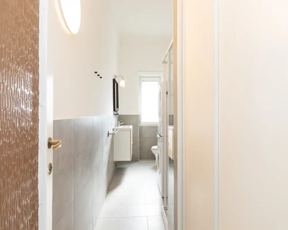 Bagno moderno e luminoso con lavabo a parete, specchio e WC in fondo; piastrelle sulle pareti inferiori e pavimento chiaro per un aspetto pulito e minimale.
