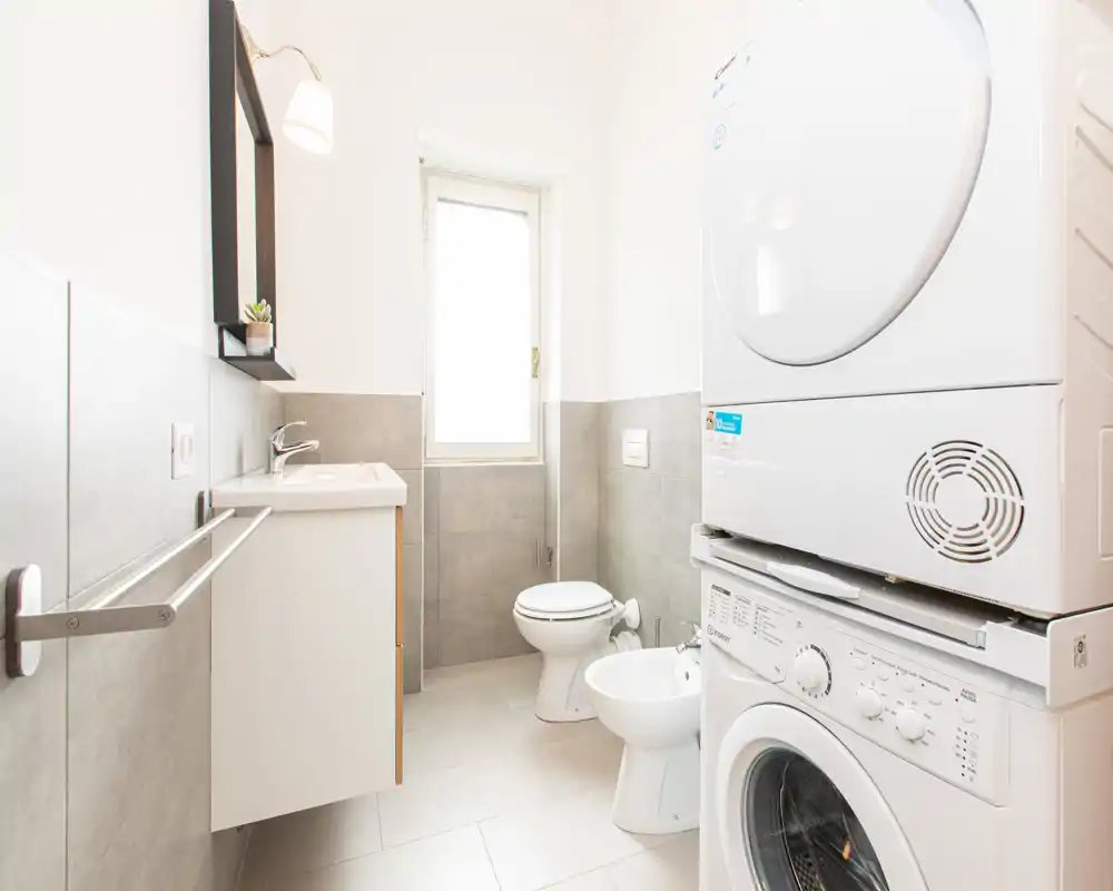 Bagno moderno e luminoso con lavatrice e asciugatrice impilate, mobile bagno sospeso, WC e bidet. La luce naturale dalla finestra mette in risalto le piastrelle pulite e i sanitari.