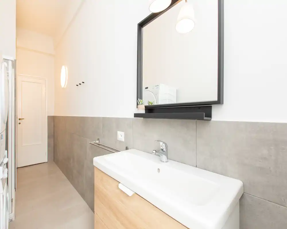 Zona lavabo bagno luminosa e moderna con grande specchio, rubinetto monocomando e mezza parete piastrellata; stile minimal e toni neutri.