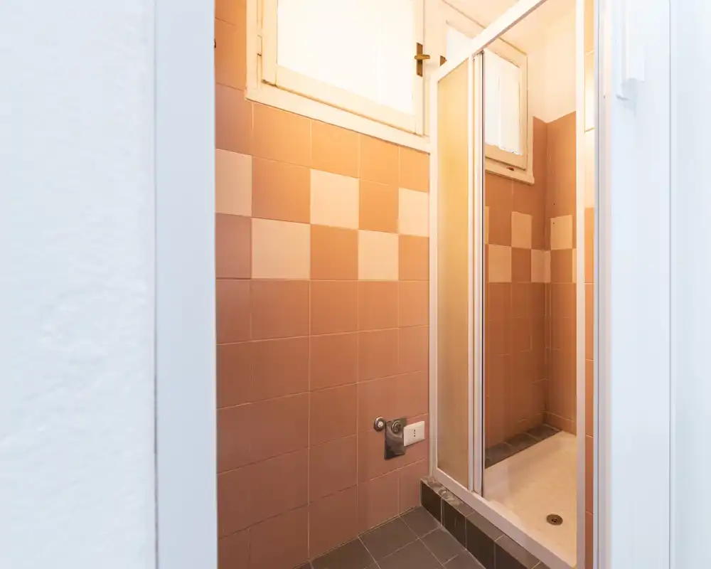 Bagno compatto con box doccia piastrellato, porta scorrevole in vetro e piastrelle pesca e crema; una piccola finestra alta garantisce luce naturale.