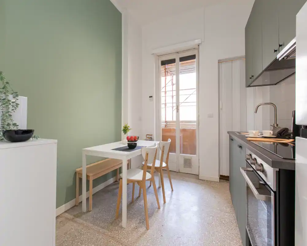 Cucina compatta e moderna con elettrodomestici integrati, tavolo da pranzo e sedie, e porta sul balcone che illumina l'ambiente con luce naturale.