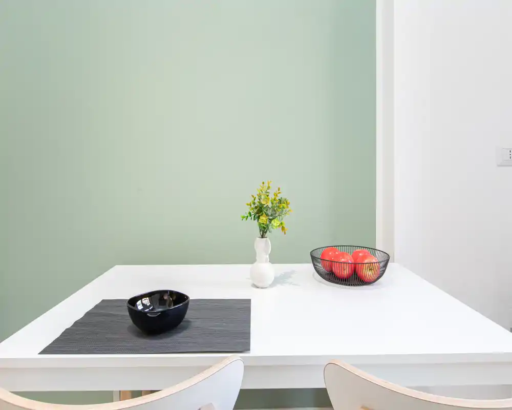 Zona pranzo luminosa e minimal con tavolo bianco pulito, vaso decorativo e ciotola di frutta — perfetta per un annuncio moderno.