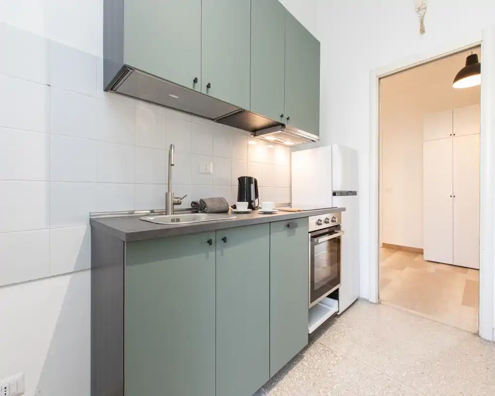 Cucina compatta e moderna con pensili verde tenue, forno integrato, lavello a incasso e piccolo frigorifero — luminosa e curata, perfetta per annunci.