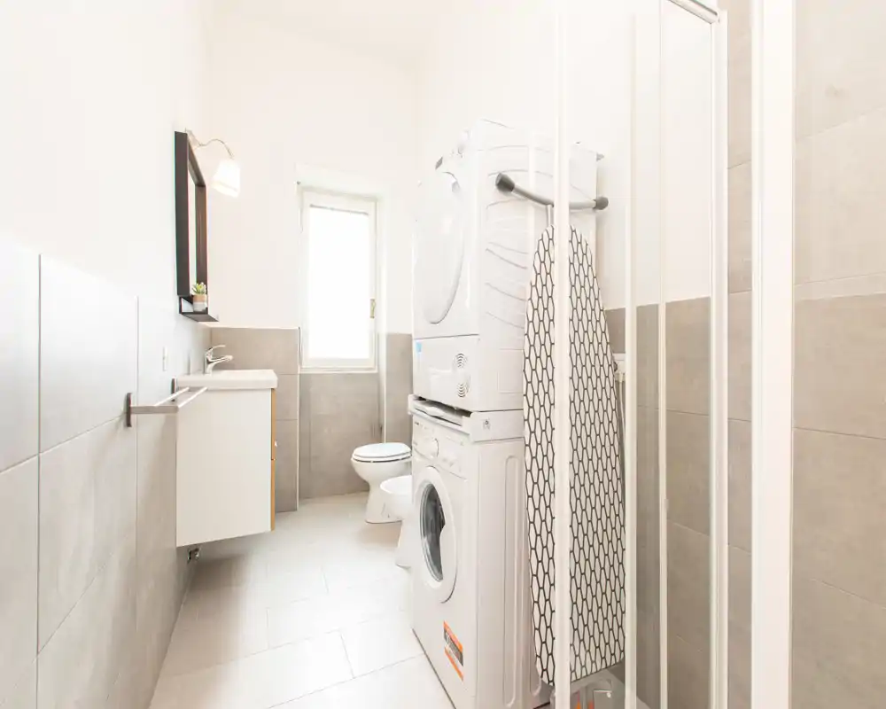 Bagno moderno e luminoso con lavatrice e asciugatrice impilate, mobile lavabo a parete, WC e doccia. Piastrelle neutre e grande finestra creano un ambiente pulito e arioso.