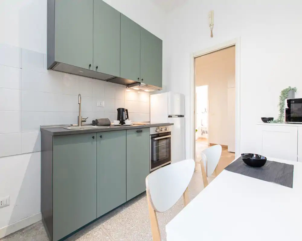 Cucina moderna e luminosa con ante verdi opache, forno incassato, frigorifero e un piccolo angolo pranzo — perfetta per appartamenti compatti.