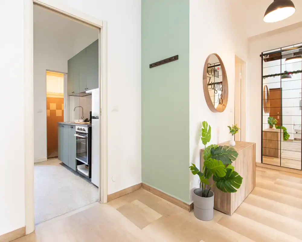 Ingresso luminoso e ordinato con uno specchio rotondo decorativo, mobile console e pianta in vaso; una porta conduce alla cucina e una parete con specchio riflette l'ambiente.