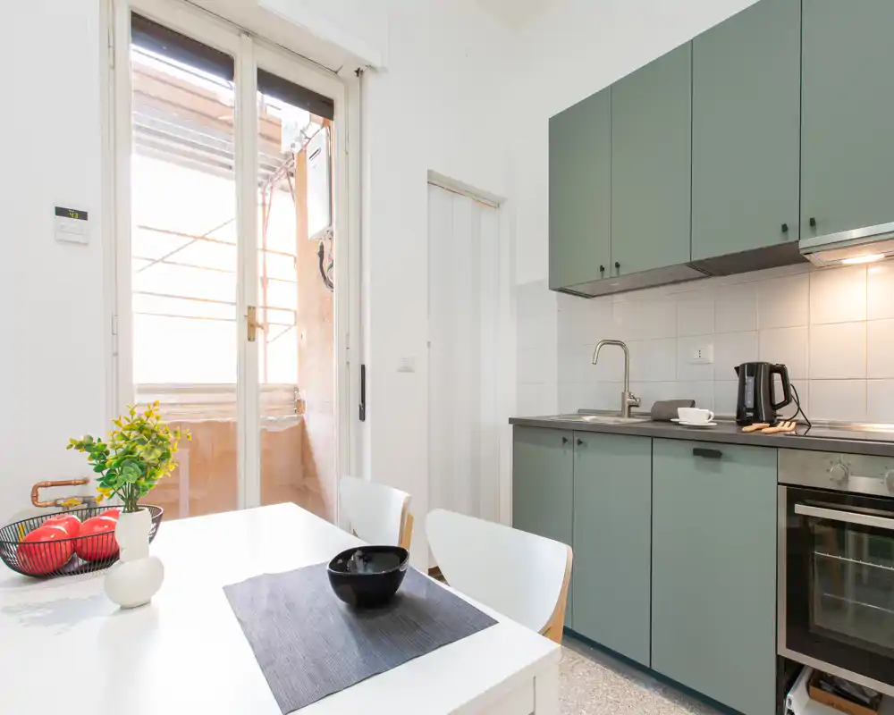 Cucina moderna compatta con mobili verde salvia, forno integrato e piccolo tavolo da pranzo accanto a una grande porta finestra che illumina l’ambiente.