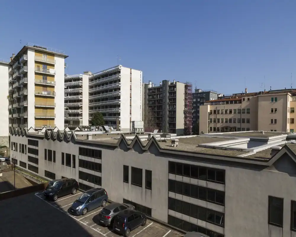 Vista exterior urbana con tejados de edificios, bloques de apartamentos y un pequeño aparcamiento bajo un cielo azul claro.