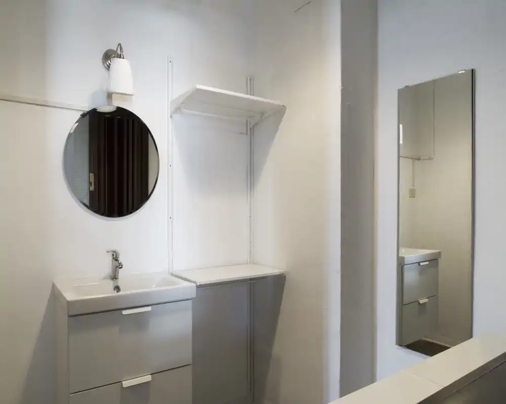 Pequeño rincón de baño moderno con mueble-lavabo, espejo circular en la pared y un espejo rectangular que refleja el mueble; paredes blancas limpias y estantería minimalista.