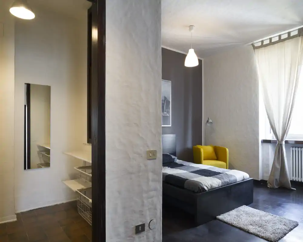 Dormitorio moderno y luminoso con cama doble, alfombra suave y un sillón amarillo junto a la ventana que deja entrar luz natural.