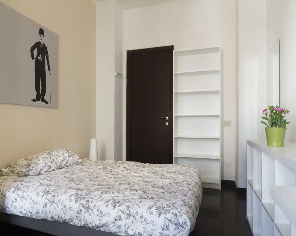 Camera da letto luminosa e ordinata con letto matrimoniale con lenzuola a motivo, scaffalatura minimal e una pianta decorativa—ambiente tranquillo e accogliente.