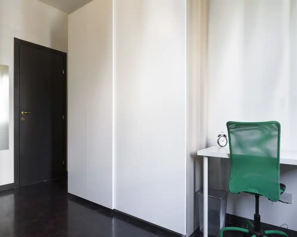 Angolo di stanza moderno e minimal con un grande armadio bianco e una piccola scrivania bianca con sedia da ufficio verde; ambiente luminoso, pulito e ordinato, adatto come area studio o per una camera compatta.