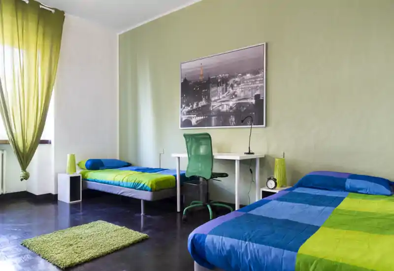 Camera da letto luminosa e moderna con due letti singoli, una scrivania e biancheria colorata. La luce naturale dalla finestra e l'arredamento verde-blu creano un ambiente fresco e accogliente.