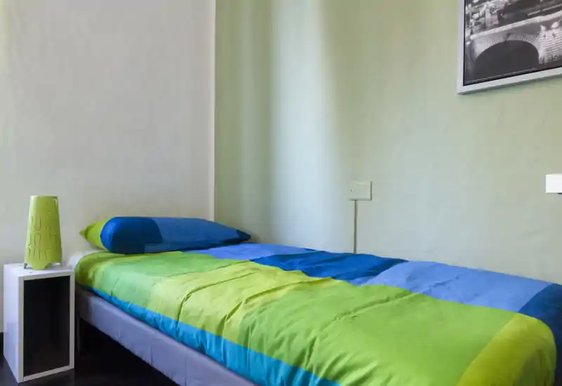 Piccola camera singola con letto rifinito in biancheria verde e blu brillante, un semplice comodino e luce soffusa e uniforme.