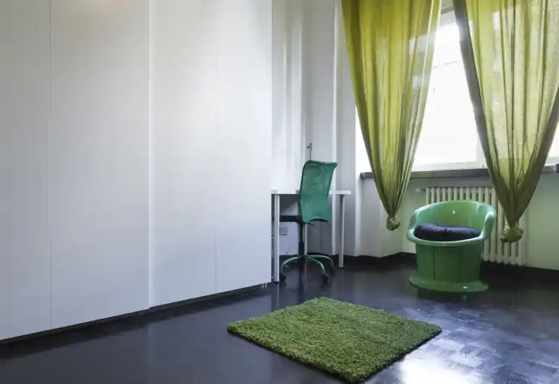 Ambiente luminoso e minimale in stile camera da letto con grande armadio bianco, una piccola scrivania e una poltrona verde; tappeto e tende verdi aggiungono colore e calore.
