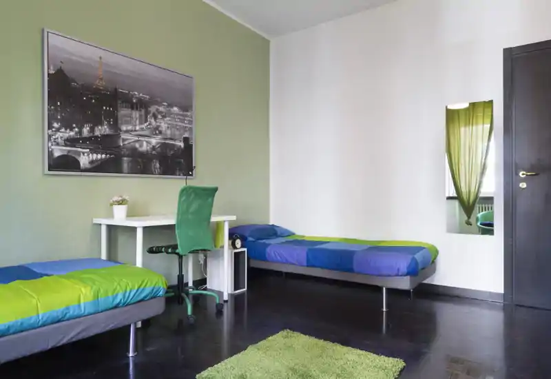 Camera luminosa e ordinata con due letti singoli, una scrivania e dettagli verdi coordinati. Arredamento moderno, sedia confortevole e una foto decorativa: perfetta per convivenze o studenti.