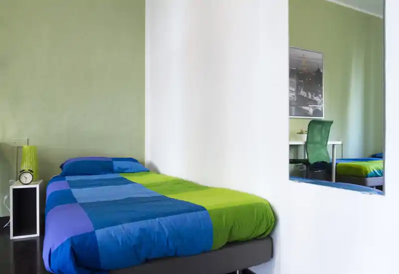 Camera da letto luminosa e moderna con una coperta blu e verde vivace, un piccolo comodino e uno specchio a parete che dà profondità alla stanza.