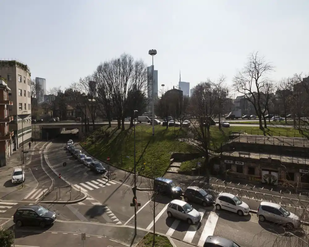 Veduta esterna urbana di un incrocio con auto parcheggiate, un terrapieno erboso e grattacieli sullo sfondo sotto luce diurna chiara.