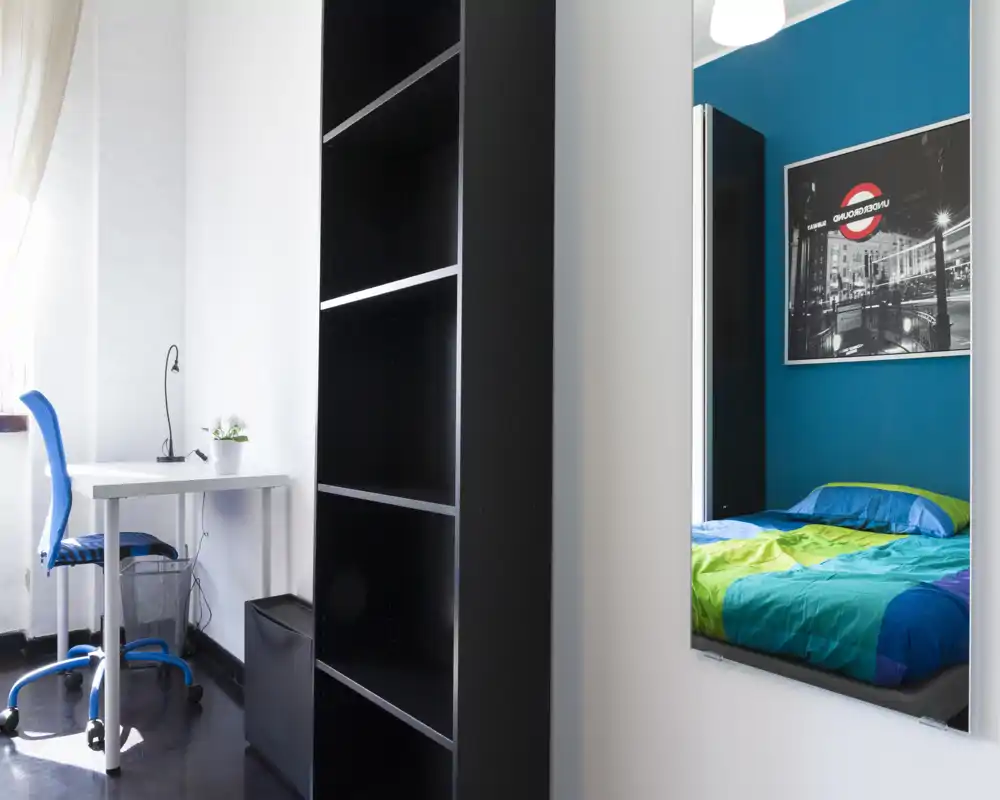 Angolo camera luminoso e moderno con biancheria colorata visibile nello specchio e una piccola area studio vicino alla finestra. L'immagine mette in evidenza i tessuti del letto e una scrivania ordinata, ideale per annunci di affitto.