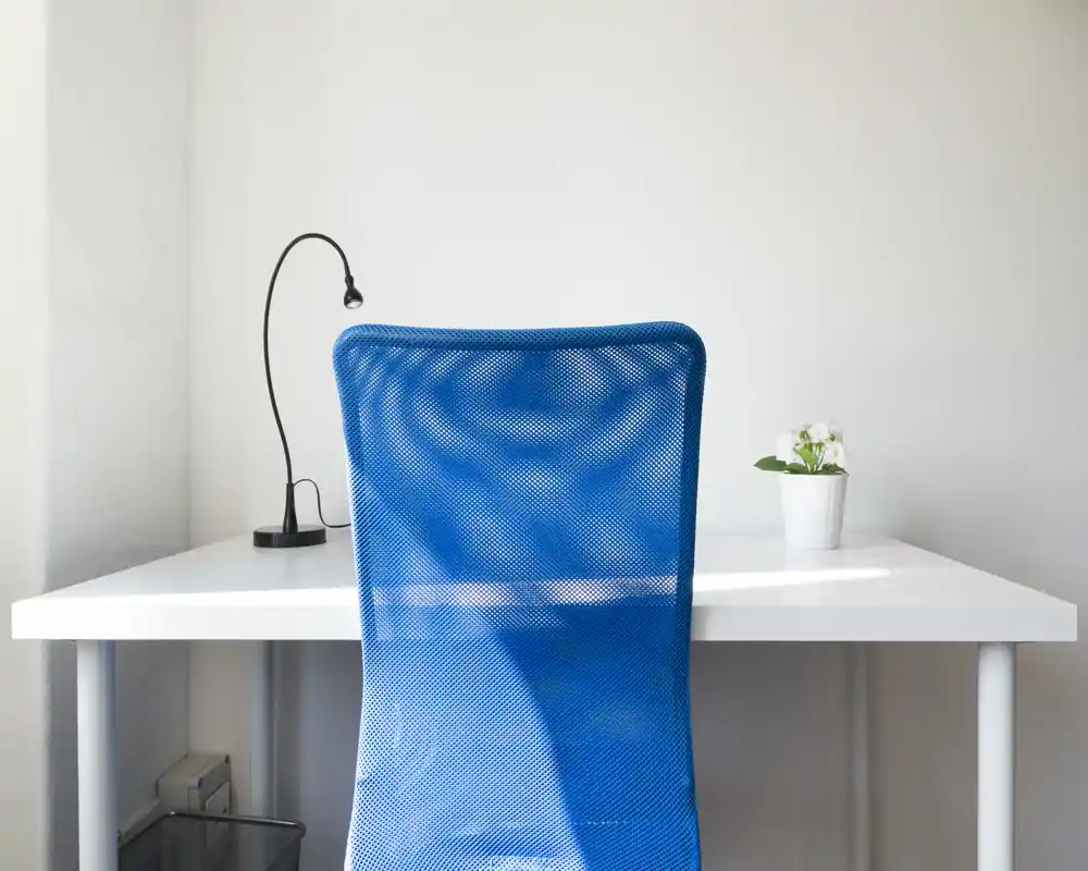 Angolo lavoro minimal con scrivania bianca, sedia da ufficio blu in rete, lampada e una piccola pianta — luminoso, semplice e funzionale.