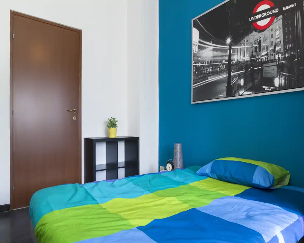 Camera da letto moderna e luminosa con parete d'accento blu e letto matrimoniale con lenzuola colorate blu e verde; scaffale semplice, lampada da comodino e poster in bianco e nero completano l'ambiente.