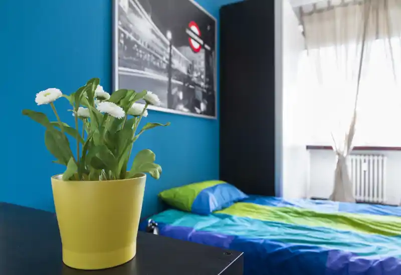 Camera da letto luminosa e colorata con letto matrimoniale con biancheria blu e verde, una stampa incorniciata e luce naturale da una finestra con tende. Un vaso giallo con fiori bianchi in primo piano aggiunge un tocco decorativo fresco.