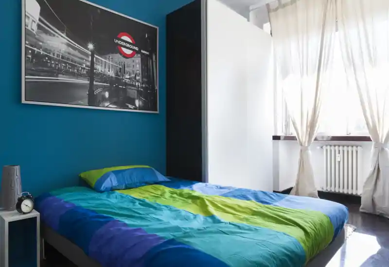 Camera da letto luminosa e colorata con letto matrimoniale, parete d'accento turchese, ampia finestra che lascia entrare molta luce naturale; armadio moderno e quadro completano l'aspetto accogliente e contemporaneo.