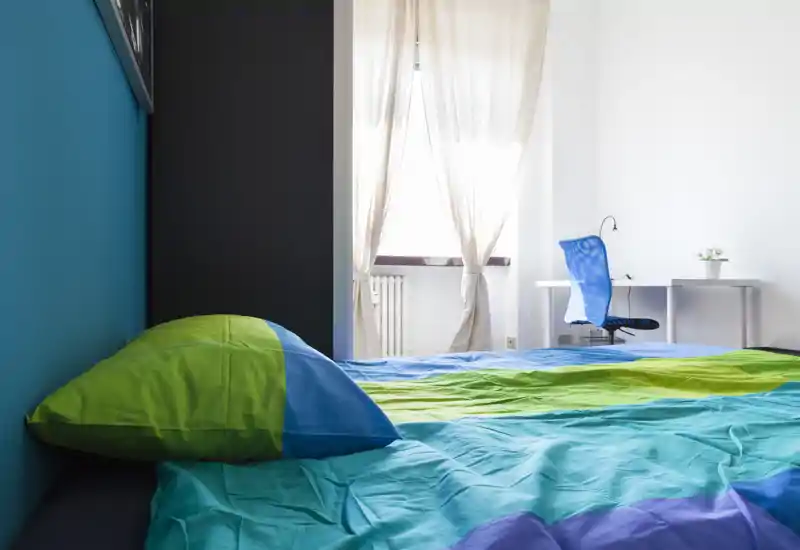 Camera da letto luminosa e moderna con biancheria colorata in primo piano e una scrivania vicino alla finestra; luce naturale e design minimale adatti per annunci di affitto.