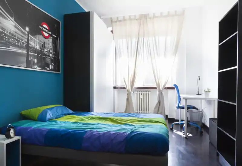 Camera da letto luminosa e moderna con letto matrimoniale con biancheria colorata, scrivania vicino alla finestra e pratici armadi. La grande finestra con tende leggere filtra la luce naturale donando un’atmosfera fresca e accogliente.