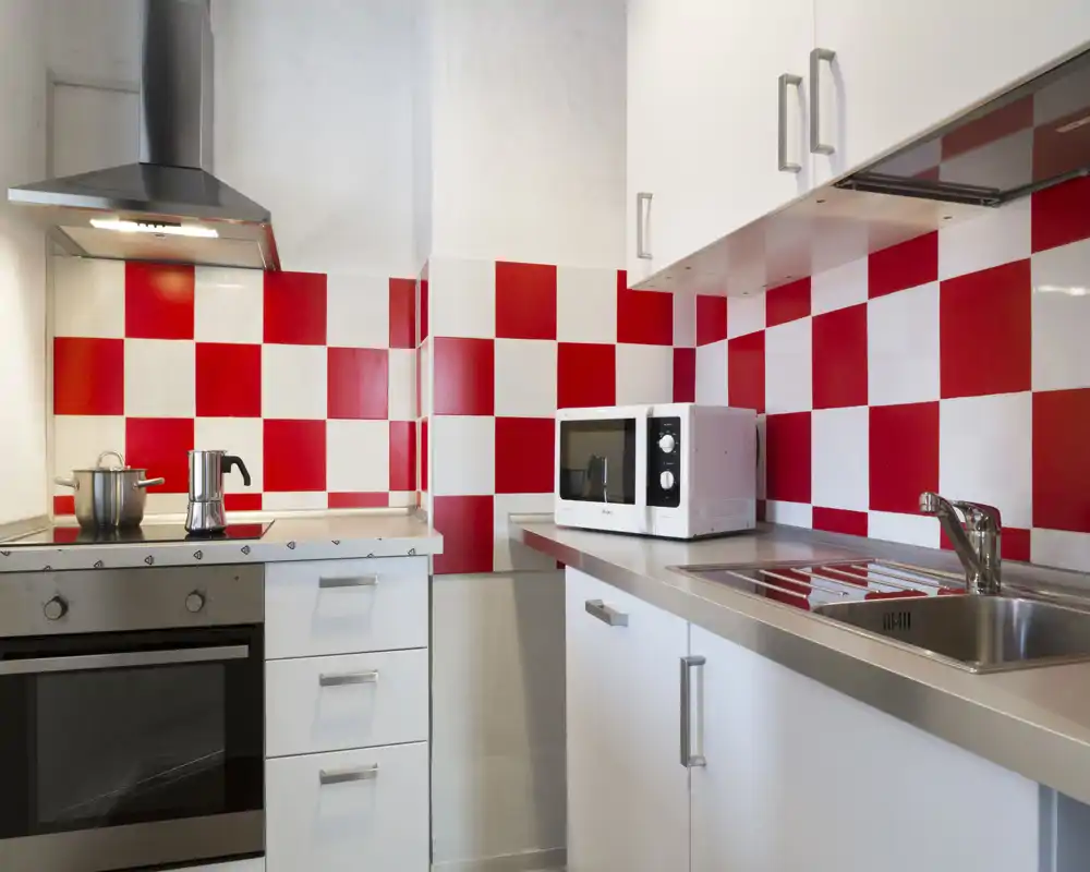 Cucina moderna compatta con piano in acciaio, forno, piano cottura e microonde; il paraschizzi a scacchi rosso e bianco dona un tocco vivace.