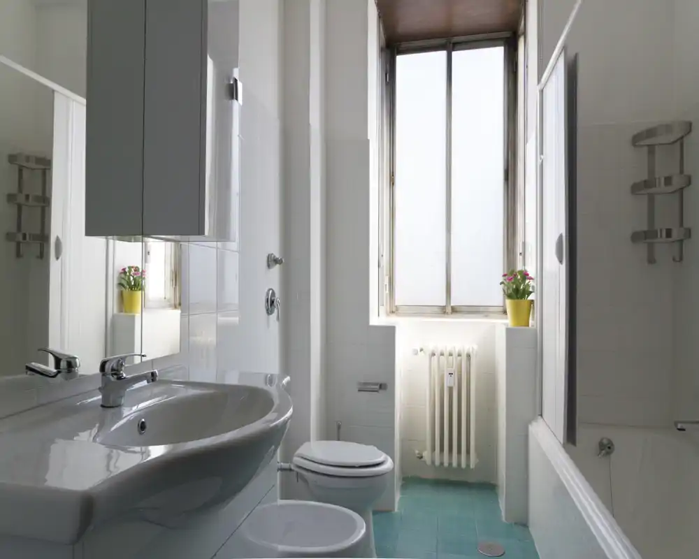 Bagno luminoso e pulito con lavabo, WC, bidet e vasca; finestra alta con vetro satinato dà luce naturale morbida e una pianta in vaso aggiunge colore.