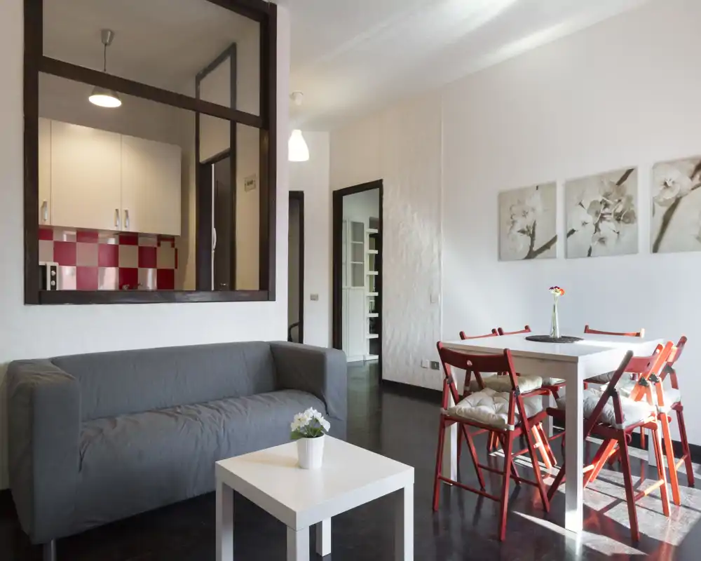 Zona giorno luminosa e ben arredata con un divano grigio, un piccolo tavolino bianco e un tavolo da pranzo con sedie rosse; il finestrino che comunica con la cucina aumenta la sensazione di spazio.