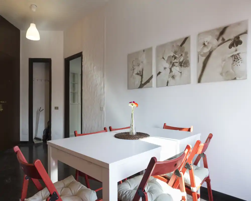 Zona pranzo luminosa e curata con tavolo bianco, sedie pieghevoli rosse e quadri floreali, perfetta per pasti informali.