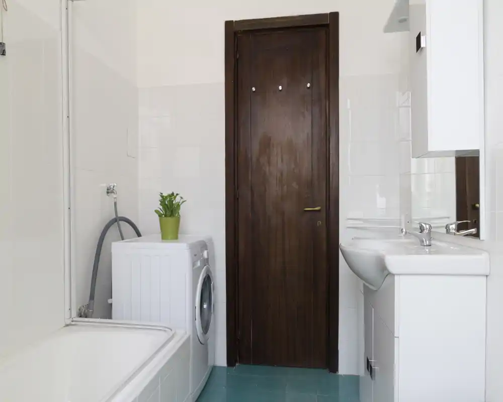 Bagno luminoso e pulito con vasca, lavabo e lavatrice accanto a una porta in legno; piastrelle bianche e una disposizione compatta ideale per soggiorni pratici.