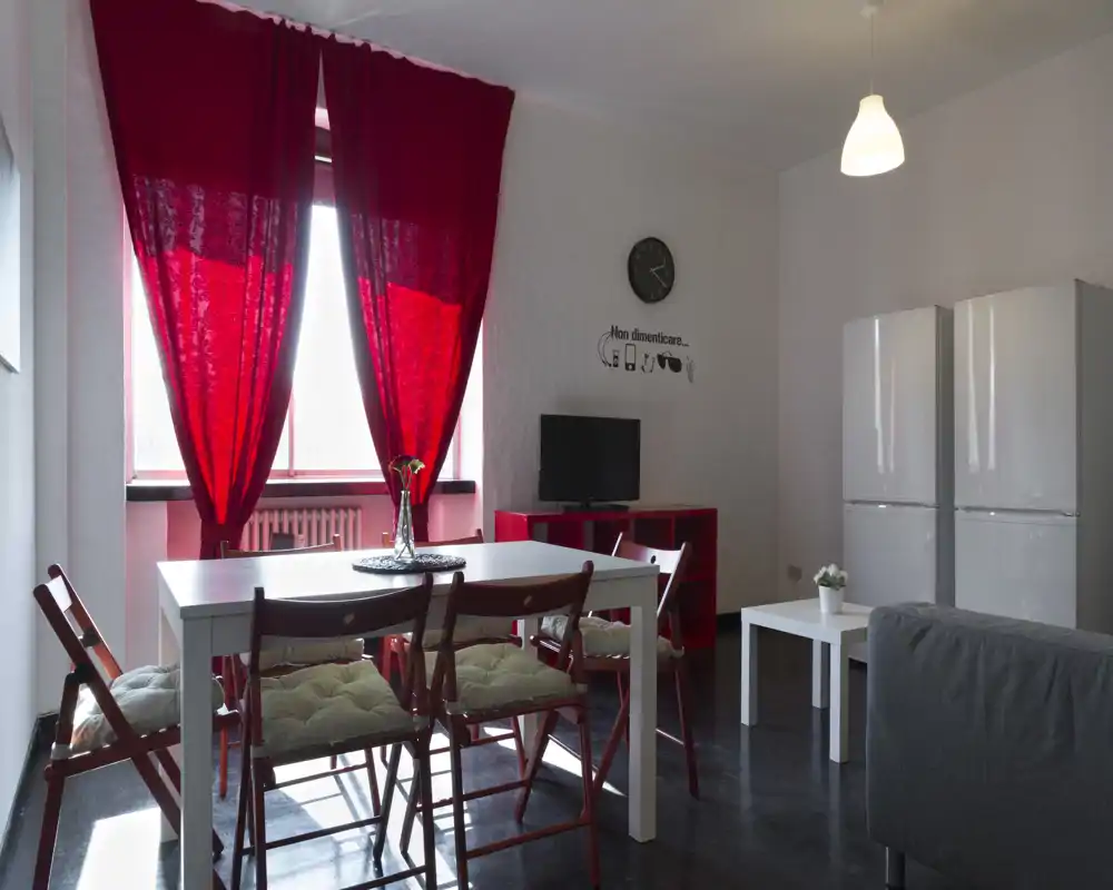 Zona giorno moderna e luminosa con tocchi rossi: tavolo da pranzo bianco con sedie pieghevoli imbottite, tende rosse, mobile TV e area salotto. Lo spazio è curato e illuminato da una finestra ampia.