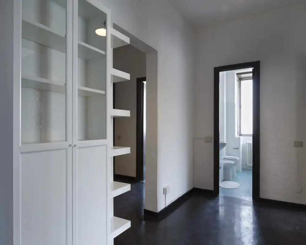 Ingresso/corridoio con scaffalatura integrata e porta visibile verso il bagno. Spazio pulito e minimalista, pavimento scuro e pareti bianche per un aspetto semplice e moderno.
